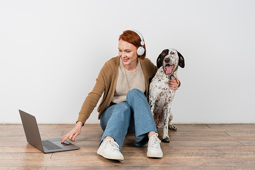 Smiling redhead woman in headphones using laptop and hugging dalmatian dog at home