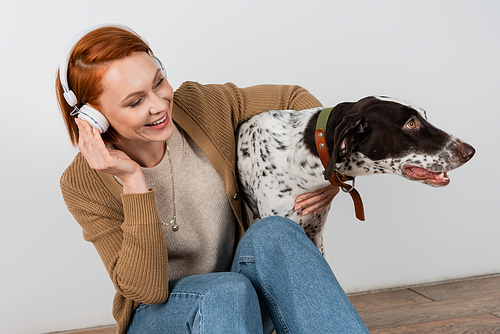 Cheerful redhead woman listening music in headphones and hugging dalmatian dog at home
