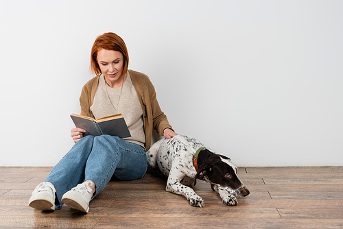 Redhead woman reading book near dalmatian dog on floor at home