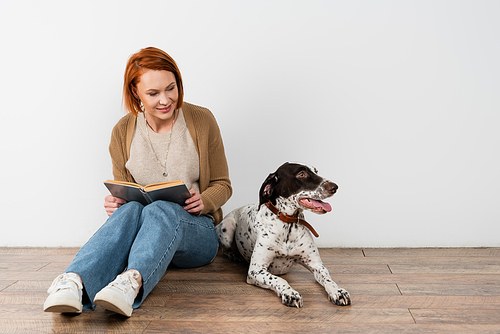 Red haired woman holding book and looking at dalmatian dog on floor at home