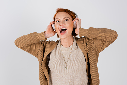 excited and red haired woman in headphones singing isolated on grey