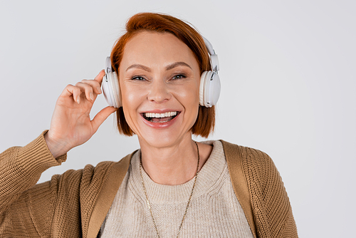 Portrait of cheerful red haired woman listening music in headphones isolated on grey