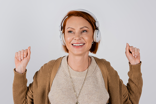 Positive red haired woman in headphones and casual clothes isolated on grey