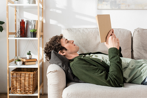 side view of young man with curly hair reading book while lying on couch in living room