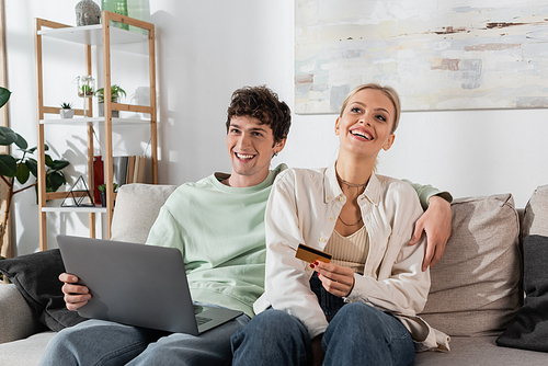 cheerful woman holding credit card near boyfriend with laptop doing online shopping