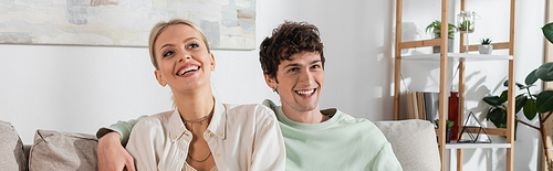 happy and curly young man hugging blonde girlfriend at home, banner