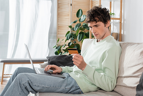 focused young man holding credit card while doing online shopping on laptop