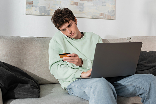 curly young man holding credit card while talking on smartphone near laptop