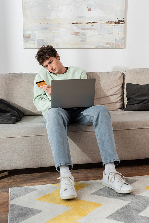 curly young man holding credit card while making order and talking on smartphone near laptop