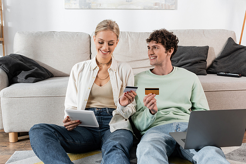 cheerful man and woman holding gadgets and credit cards while doing online shopping