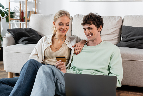 cheerful man holding credit card while using laptop near joyful girlfriend