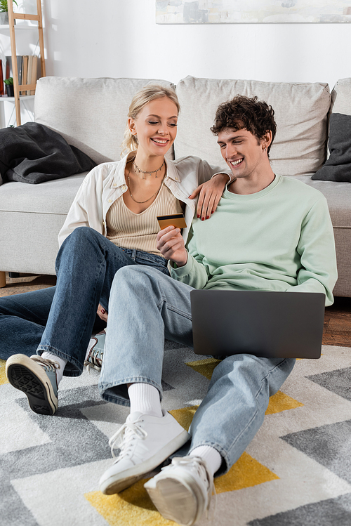 cheerful man holding credit card while using laptop near smiling girlfriend