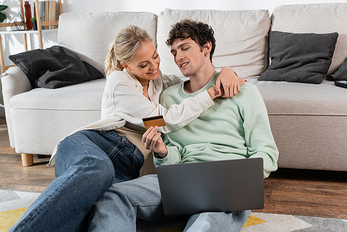 happy blonde woman hugging boyfriend holding credit card while using laptop