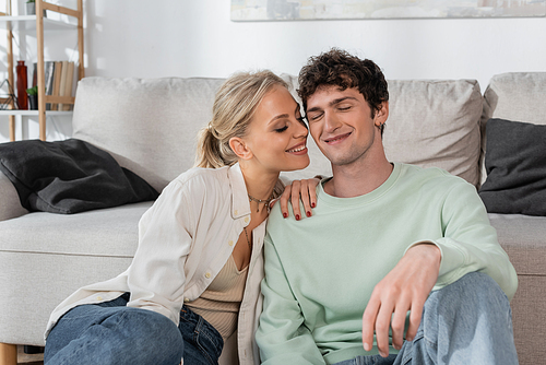 happy blonde woman with closed eyes smiling near boyfriend in living room