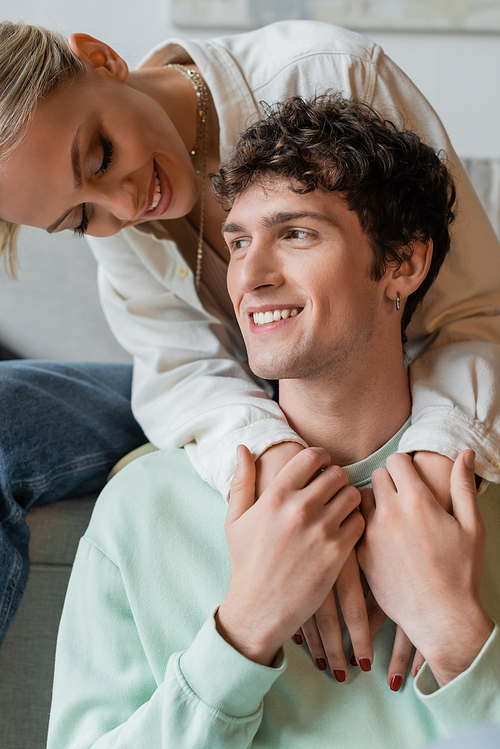 happy blonde woman embracing cheerful curly man in living room