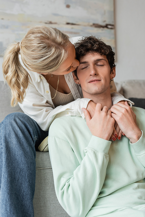 young blonde woman embracing and kissing cheek of curly man in living room