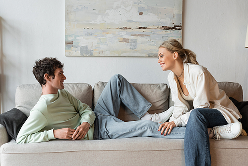 side view of cheerful young man in casual clothes and shoes resting on sofa and looking at girlfriend