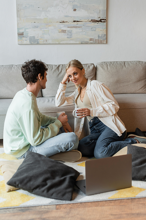 cheerful young couple holding cups of coffee near pillows and laptop on carpet