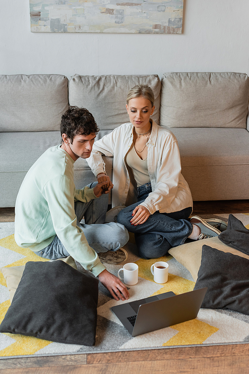curly man holding hand of blonde woman while looking at laptop near cups on carpet