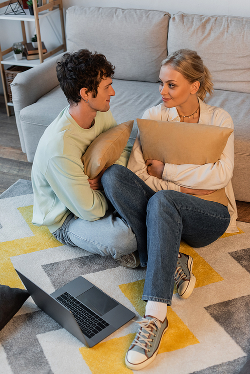 happy woman and curly man looking at each other while holding pillows near laptop on carpet
