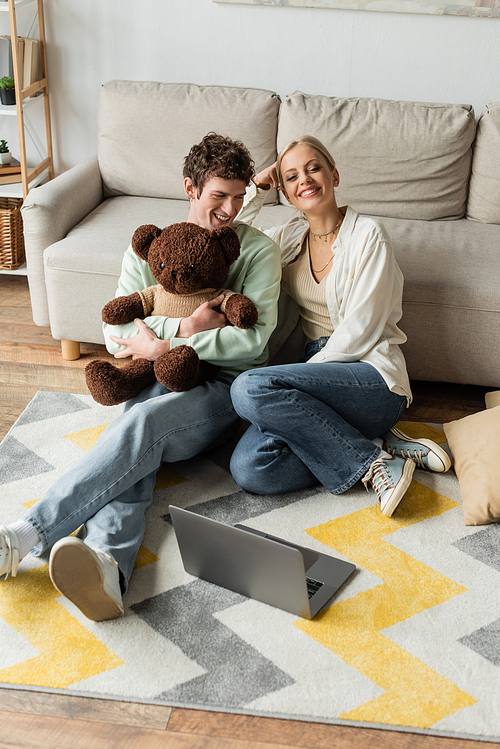 blonde woman smiling near curly young man holding teddy bear while looking at laptop on carpet