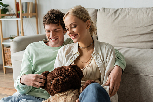 blonde woman smiling near curly young man and holding teddy bear in living room