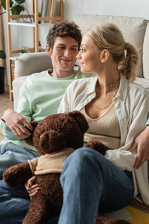 cheerful young woman and happy man holding teddy bear in living room