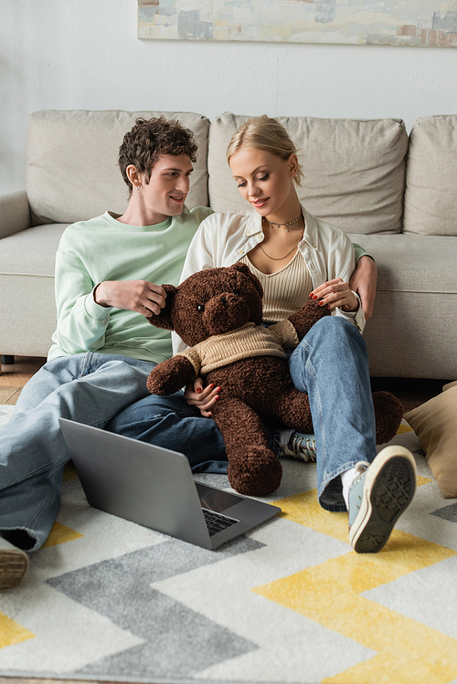 cheerful young woman and happy man holding teddy bear near laptop in living room