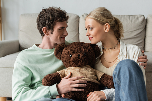 positive young woman and happy man holding teddy bear in living room