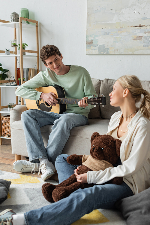 curly man sitting on couch and playing acoustic guitar near blonde woman with teddy bear