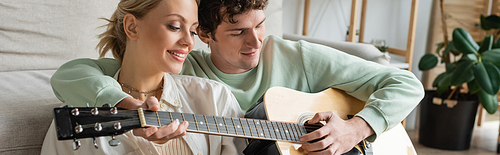 curly man playing acoustic guitar near cheerful blonde woman while sitting near couch, banner