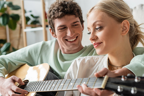 happy man playing acoustic guitar near blonde woman on blurred foreground