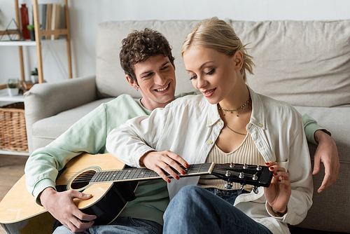 curly man playing acoustic guitar near pleased blonde woman while sitting in living room