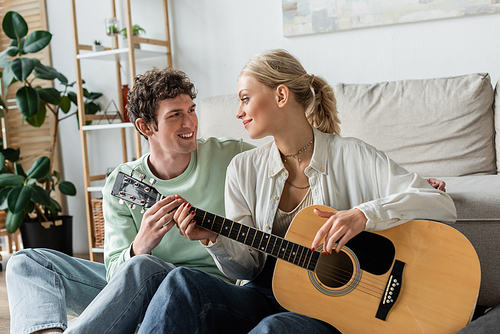 happy blonde woman playing acoustic guitar near smiling and curly boyfriend