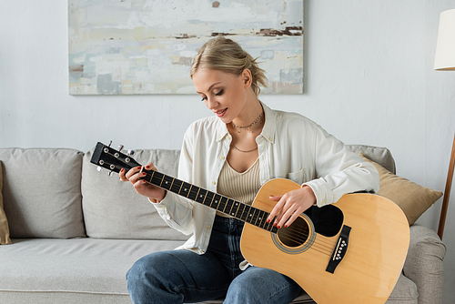 cheerful blonde woman playing acoustic guitar while sitting on couch