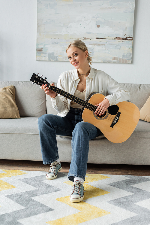 positive and blonde woman playing acoustic guitar while sitting on couch