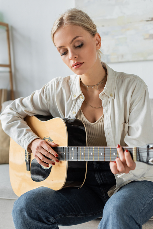 young and blonde woman playing acoustic guitar while sitting on couch