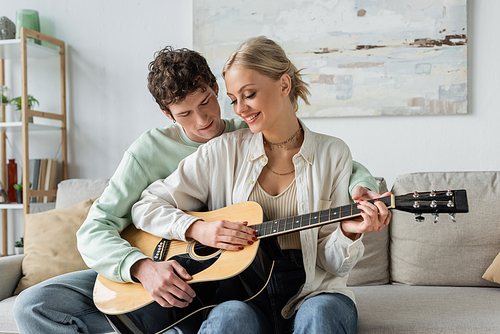 cheerful blonde woman playing acoustic guitar with curly boyfriend while sitting on couch