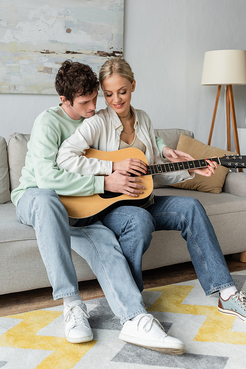 happy blonde woman playing acoustic guitar with curly boyfriend while sitting on couch