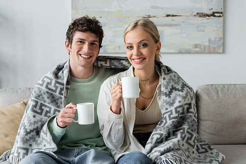 happy young man and smiling blonde woman covered in blanket holding cups of tea while sitting on couch