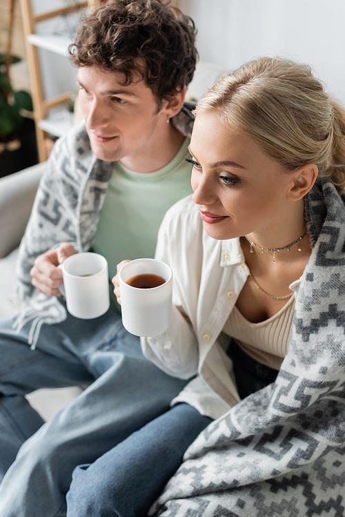 high angle view of young man and smiling woman covered in blanket holding cups of tea while sitting on couch