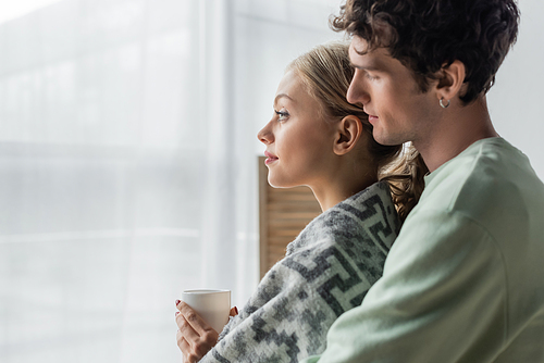 side view of caring man hugging blonde girlfriend covered in blanket and holding cup of tea