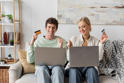 happy young couple holding credit cards near laptops while doing online shopping