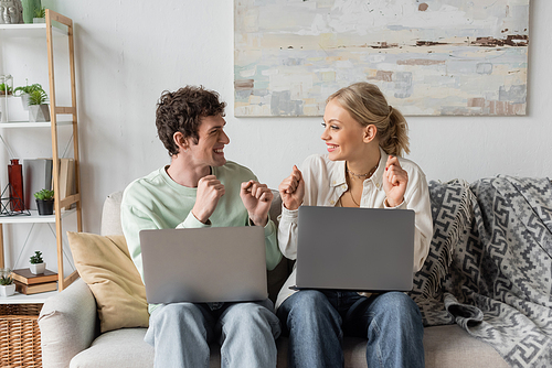 excited young bloggers rejoicing near laptops while sitting on couch