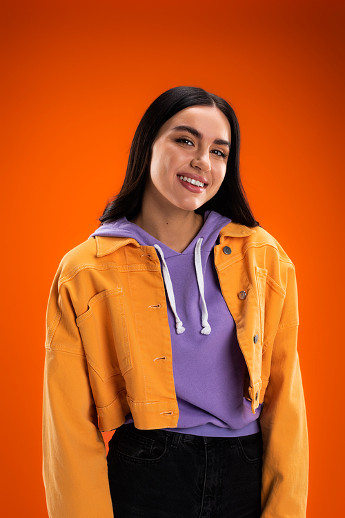 young woman in purple hoodie and stylish jacket smiling at camera isolated on orange