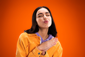 Young brunette woman pouting lips and closing eyes isolated on orange