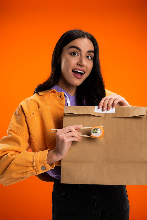 amazed woman holding chopsticks with sushi roll near paper bag isolated on orange