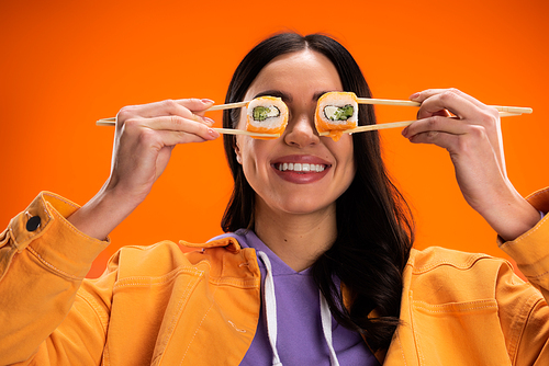cheerful woman in jacket and purple hoodie covering eyes with sushi rolls isolated on orange