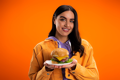 cheerful woman holding tasty burger with meat and cheese isolated on orange