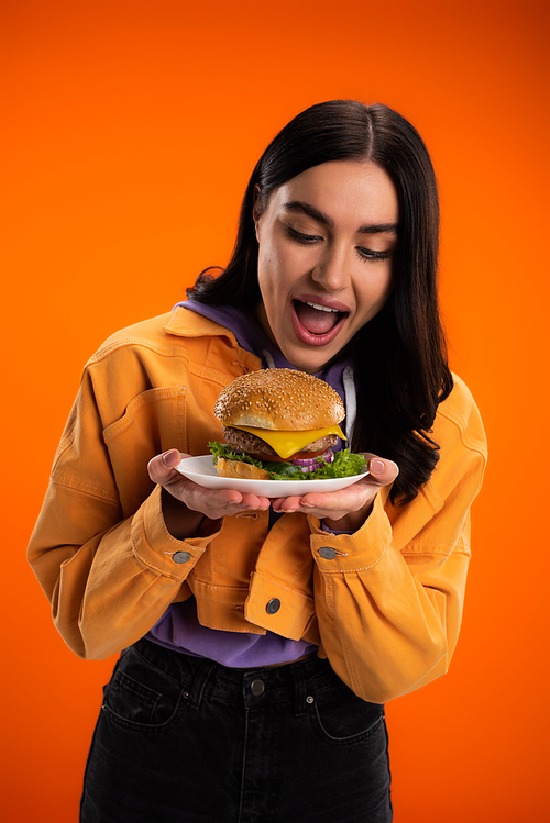 hungry woman opening mouth near plate with delicious burger isolated on orange